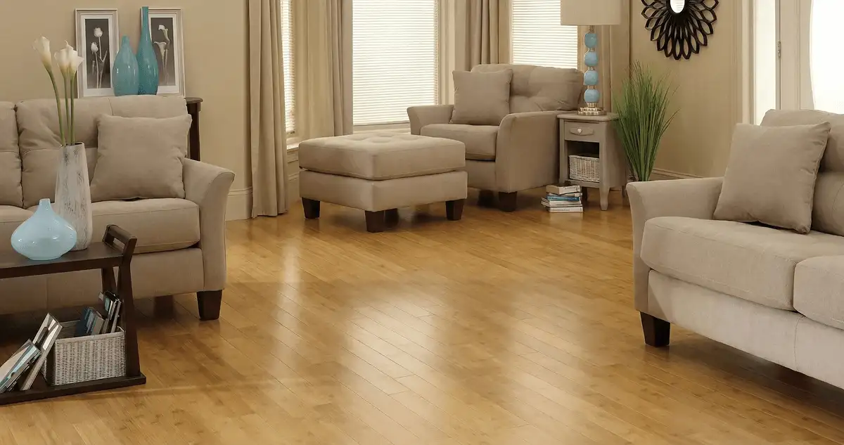 A living room with professionally installed bamboo flooring, beige sofas, a matching ottoman, a side table with a blue lamp, vases, framed pictures, and large windows with blinds and curtains in Cedar Rapids, IA